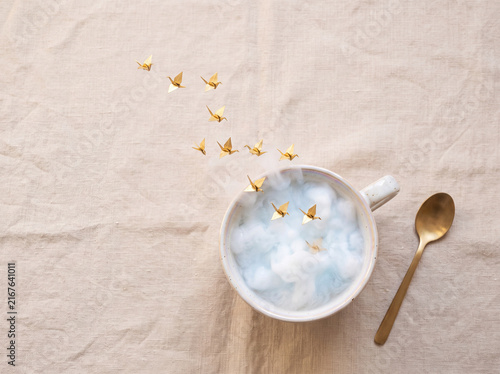 Conceptual still life of a ceramic cup filled with fluffy clouds and flying golden origami cranes on beige linen tablecloth, top view