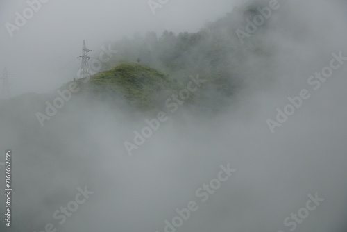 Fotografía Heavy morning mist engulfing a steep green hillside featuring a high-voltage transmission tower