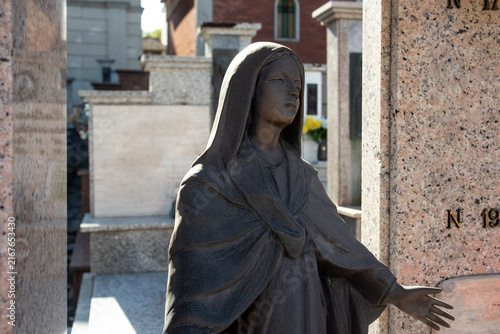 Statua in bronzo della Vergine Maria a braccia aperte posta all'ingresso di una cappella di famiglia in un cimitero, scultura sacra tra tombe e fiori