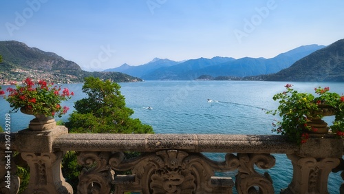 Canvas Print Scenic Lake Como view from Villa del Balbianello terrace in Italy, framed by stone balustrade, red flowers, boats and Alpine mountains