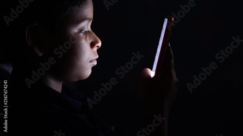 Young Indian Boy Browsing Mobile Phone At Night. Absorbed Kid Using Mobile Phone In Dark Indoors Screen Light Casting Silhouette.