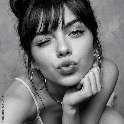 Playful black and white portrait of a beautiful young woman winking and blowing a kiss with pouty lips while resting her head on her hand.
