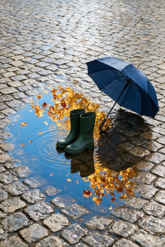 Green rubber boots and a blue umbrella on cobblestones with puddle  
