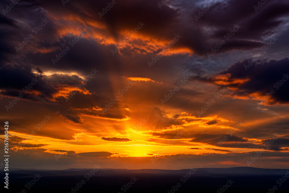 Fototapeta premium Dramatic orange sunset with sun rays through dark clouds over distant hills