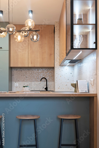 Modern kitchen interior with kitchen cabinet with glass and wood and wooden countertop with chair. New apartment with luxurious lamp and green furniture. Vertical.