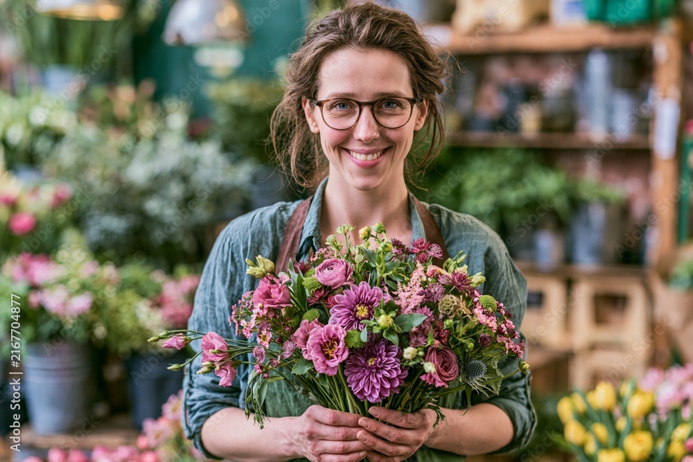 Fototapeta premium Flower Shop Owner Smiles While Holding a Mixed Flower Bouquet in a Busy Flower Shop Filled With Plants
