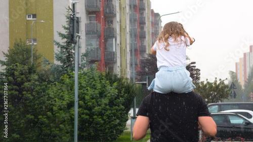 Father Carrying Little Daughter On Shoulders Running Through Heavy Summer Rainstorm Near Residential Apartment Buildings