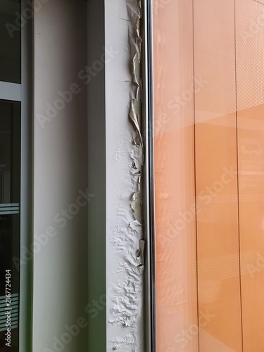 Close-up view of building facade deterioration showing severe paint peeling and water damage on a horizontal structural ledge beneath orange-tinted glass panels. The white painted surface exhibits