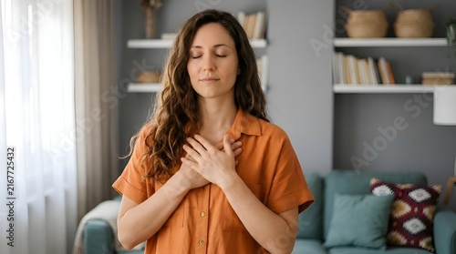 Calm adult woman with eyes closed placing hands on her chest in a peaceful moment of gratitude and mindfulness inside a bright modern living room with soft natural indoor lighting effects.