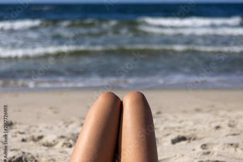 Woman's Tanned Legs Relaxing on Sandy Beach Facing Ocean Waves