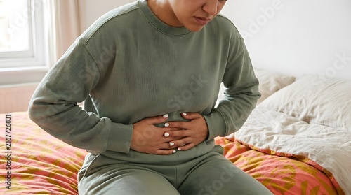 Woman in light green loungewear sitting on a bed while holding her stomach in pain due to abdominal cramps or indigestion in a bright indoor room during the daytime with soft lighting