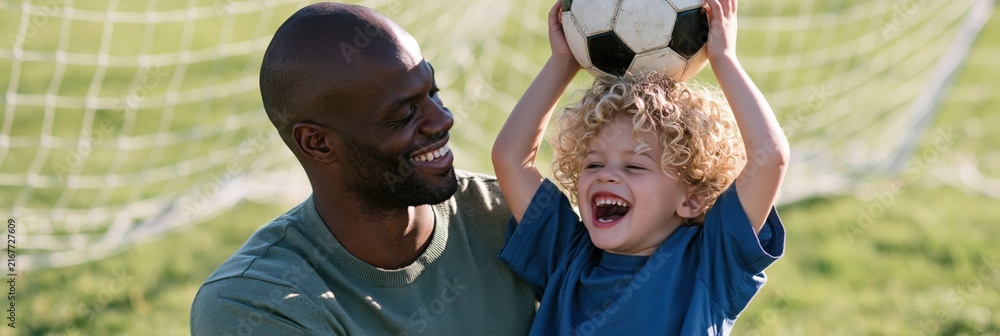 Obraz premium Smiling father watching son hold a soccer ball above his head by the goal on grassy field outdoors for family sports banner poster background and lifestyle design