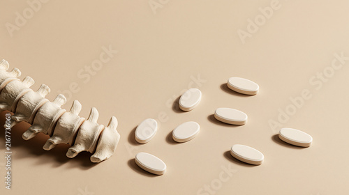 Exclusive photo of Human spine model beside oval white pills on a beige surface. Represents pain management and spinal health treatment solutions.