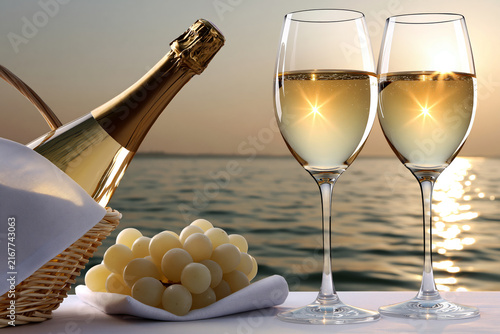 Two glasses of champagne with sunset reflection beside picnic basket and grapes by the water at golden hour