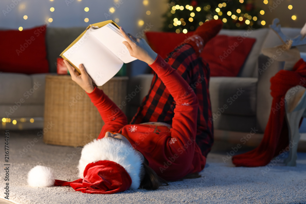 Fototapeta premium Young girl in Santa hat reading book at home on Christmas eve