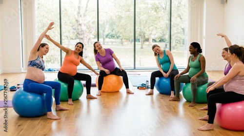 Group of pregnant women in colorful athletic wear balancing on large exercise balls in bright fitness studio with big windows