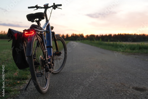 Electric bicycle with illuminated rear light and pannier bag parked on a rural road at sunset, with open countryside, warm evening sky and ample copy space on the right