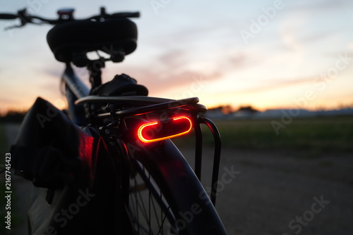 Electric bicycle with illuminated red rear light, rear rack and pannier bag on a rural path at sunset, with blurred background and copy space on the right