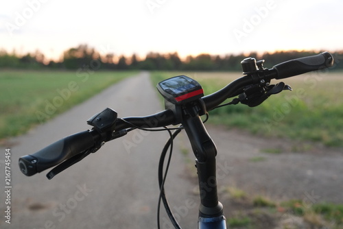Electric bicycle handlebars and display seen in close-up on a quiet rural path at sunset, with green fields, soft evening light, blurred background and generous copy space