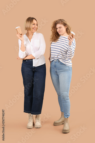 Beautiful happy women in stylish jeans with cups of coffee on beige background
