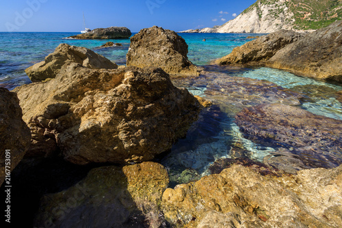 A photograph of the beautiful, azure Paralia Petani beach with large stones in the foreground.