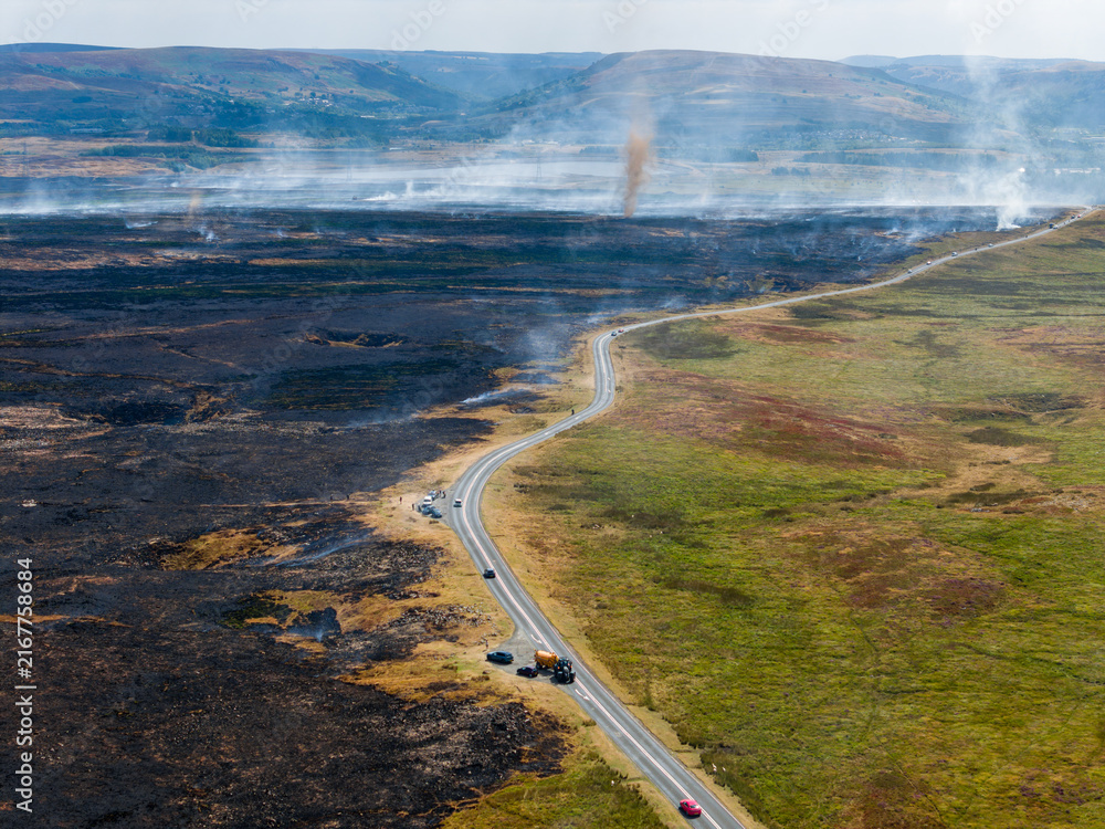 Obraz premium Smoke whirl forming above burnt heathland after summer wildfire, vehicles parked along road dividing charred landscape from green moor, South Wales