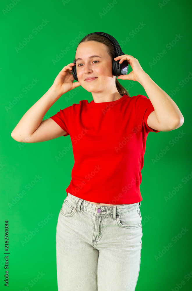 Fototapeta premium Passionate teenage girl in red T-shirt and jeans listening and dancing to music on headphones on a green background. Chroma key compositing