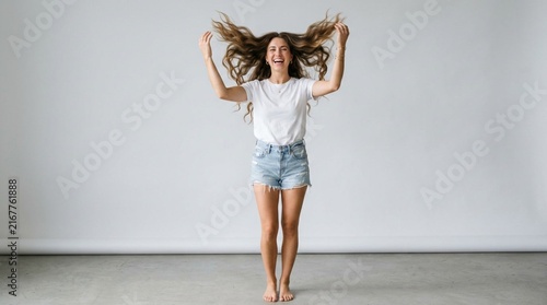 Joyful young woman laughing and tossing long wavy hair with both hands, wearing a casual white t-shirt and denim shorts, barefoot full length studio portrait against light gray wall, happy lifestyle