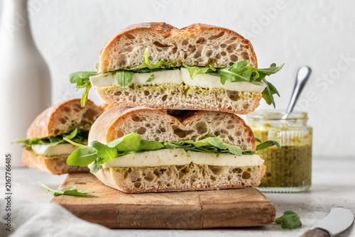 Fresh ciabatta sandwiches with mozzarella, arugula and pesto sauce in stack on wooden board, toasted italian panini, rustic deli