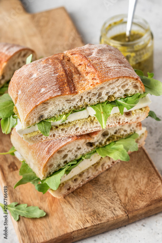 Fresh ciabatta sandwiches with mozzarella, arugula and pesto sauce in stack on wooden board, toasted italian panini, rustic deli