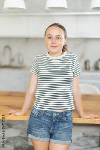 Teenage girl stands in a bright kitchen near a wooden table and thinks about what to cook for breakfast