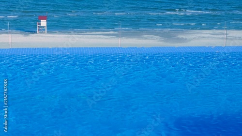 Swimming pool water rippling with ocean beach background. Crystal clear blue water of a swimming pool gently rippling with a sandy beach and the calm sea in the background, creating a serene