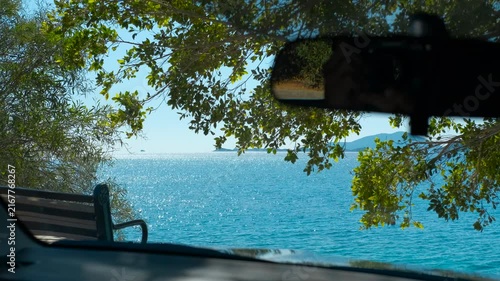 Vehicle parked near coast enjoying stunning ocean view. Sunlight reflects on a calm, blue ocean, seen through the windshield of a parked vehicle near a leafy tree and a bench, offering a coastal view