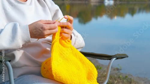 Woman knitting with yellow yarn outdoors by a lake. Calm woman sitting on a camping chair and knitting with bright yellow yarn, enjoying a peaceful hobby in nature by the tranquil water