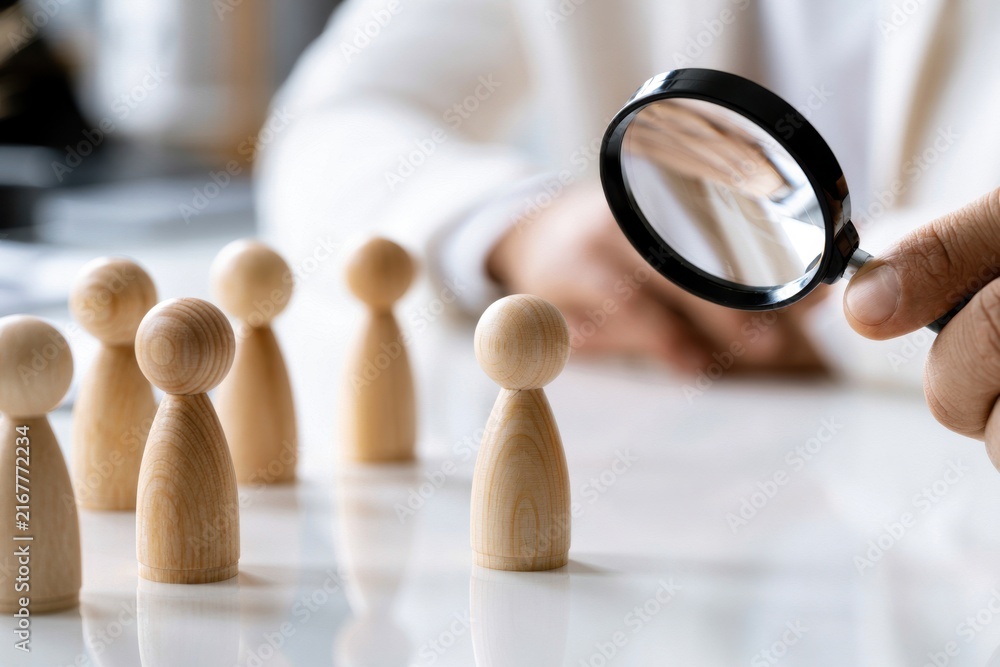 Fototapeta premium Person examining one wooden figure among a group with a magnifying glass representing selection and detailed evaluation in recruitment or strategy planning