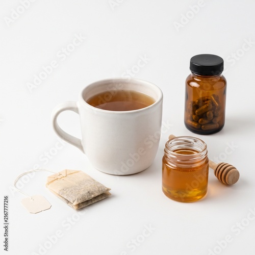 A cup of hot herbal tea with a tea bag, a small jar of honey, and a bottle of pills on a white surface. The tea is steeping and emitting steam, suggesting a soothing and healing beverage.