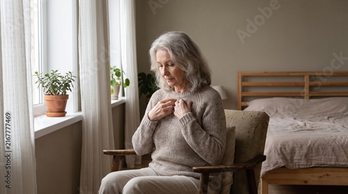 Elderly Woman Sitting In Armchair By Window Holding Chest With Concerned Expression