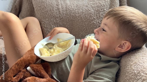 Happy little boy lying down back and eating delicious homemade dumplings with sour cream, relaxed cute child enjoying food on cozy sofa