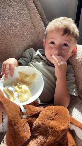 Happy little boy lying down back and eating delicious homemade dumplings with sour cream, relaxed cute child enjoying food on cozy sofa