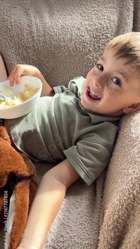 Happy little boy lying down back and eating delicious homemade dumplings with sour cream, relaxed cute child enjoying food on cozy sofa