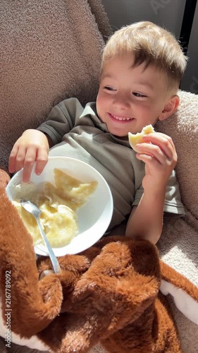 Happy little boy lying down back and eating delicious homemade dumplings with sour cream, relaxed cute child enjoying food on cozy sofa
