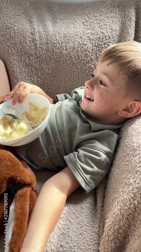 A cheerful three year old toddler mischievous smiling on camera while eating homemade cheese dumplings in a cozy and soft armchair.
