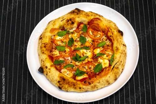 Neapolitan-style personal pizza with naturally leavened long-fermentation dough, sliced tomatoes, melted mozzarella, fresh basil and oregano, served on a white plate over a dark pinstriped cloth.