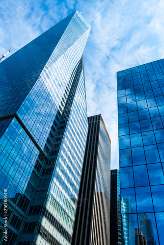Modern skyscrapers and glass office buildings rise above an urban business district, featuring reflective facades, dramatic perspective, blue sky, and contemporary city architecture.