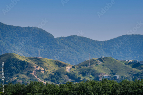 Scenic vista of rolling green grassy hills with dirt roads and communication towers set against a backdrop of dense forested mountain ranges under a clear sky