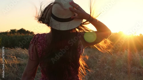 Woman from behind with long hair and a hat walking through a golden field of tall grass during a beautiful sunset, with the wind blowing through her hair and warm lens flare
