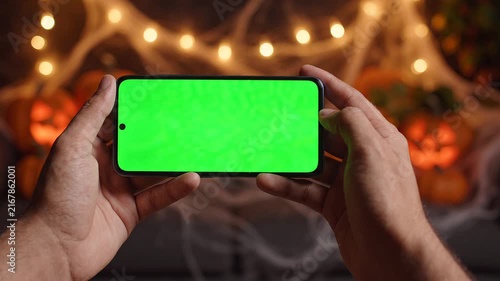 Person Holding a Smartphone with a Green Screen Chroma Key Display in Front of Blurred Halloween Pumpkin Decorations
