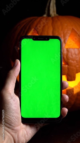 Person Holding a Smartphone with a Green Screen Chroma Key Display in Front of a Glowing Jack O Lantern Halloween Pumpkin