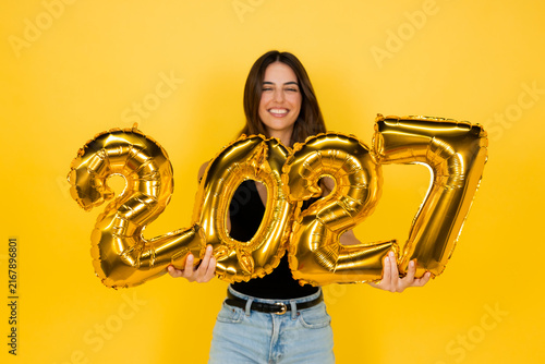 Young woman smiling while holding shiny gold foil balloons forming the numbers 2027 against a vibrant yellow background, celebrating the upcoming new year