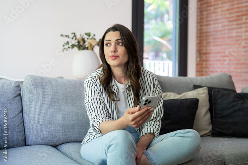 Foto Woman sitting cross-legged on a gray couch, holding a smartphone, looking away w